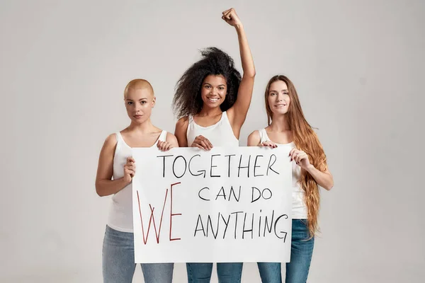 Tre självsäkra olika kvinnor i vit skjorta och jeans ser glada ut när de håller, står med en banderoll i händerna isolerade över grå bakgrund — Stockfoto