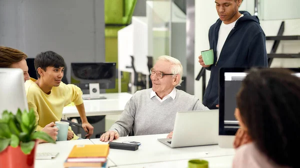 Åldrad man, seniorpraktikant som ler medan han pratar med sina unga kollegor, Vänliga arbetare som pratar med nyanställda under fikapausen på jobbet — Stockfoto