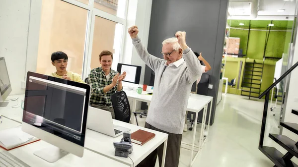 Åldrad man, seniorpraktikant ser glad ut, höjer armarna efter att ha avslutat sin första uppgift på jobbet, Vänliga arbetare applåderar, hejar på ny anställd på kontoret — Stockfoto