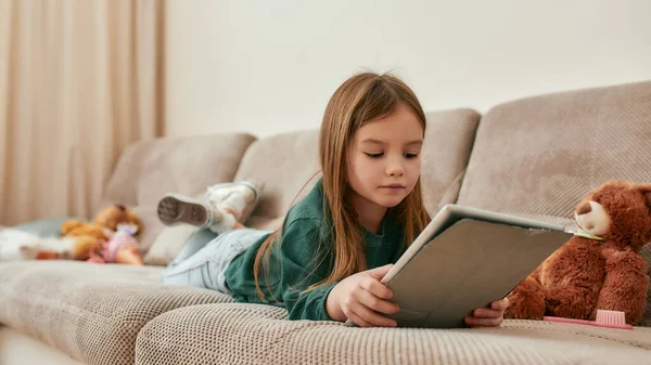 En liten flicka som ligger på magen och håller i en tablett på en soffa — Stockfoto