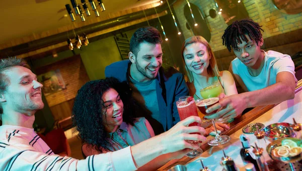 Una vida llena de celebración. Grupo de amigos, adultos jóvenes, hombres y mujeres divirtiéndose mientras brindan, bebiendo cócteles en el mostrador del bar en el club nocturno — Foto de Stock