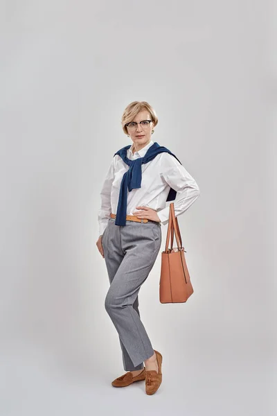 Tiro de longitud completa de mujer caucásica de mediana edad elegante con traje de negocios y gafas mirando a la cámara mientras posa con el bolso, de pie aislado sobre fondo gris —  Fotos de Stock