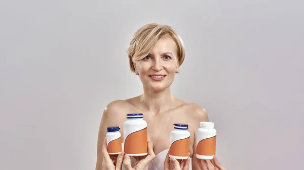 Portrait d'une femme caucasienne d'âge moyen souriant à la caméra, isolée sur fond gris. Mains tenant des bouteilles avec des pilules, des vitamines. Concept de soins de santé — Photo