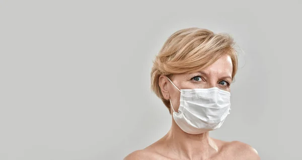 Close up portrait of beautiful middle aged woman wearing white medical mask and looking at camera isolated against grey background. Pandemic, safety, prevention concept — Stock Photo, Image