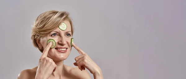 Retrato de mulher caucasiana de meia-idade atraente aplicando fatias de pepino em seu rosto como máscara, olhando para longe isolado sobre fundo cinza. Skincare, conceito de beleza — Fotografia de Stock