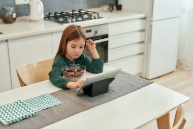 Küçük tatlı bir kız kahvaltı sırasında tabletinde ne izleyeceğini seçiyor. Uzaktan eğitim sırasında mutfakta tek başına oturuyor.