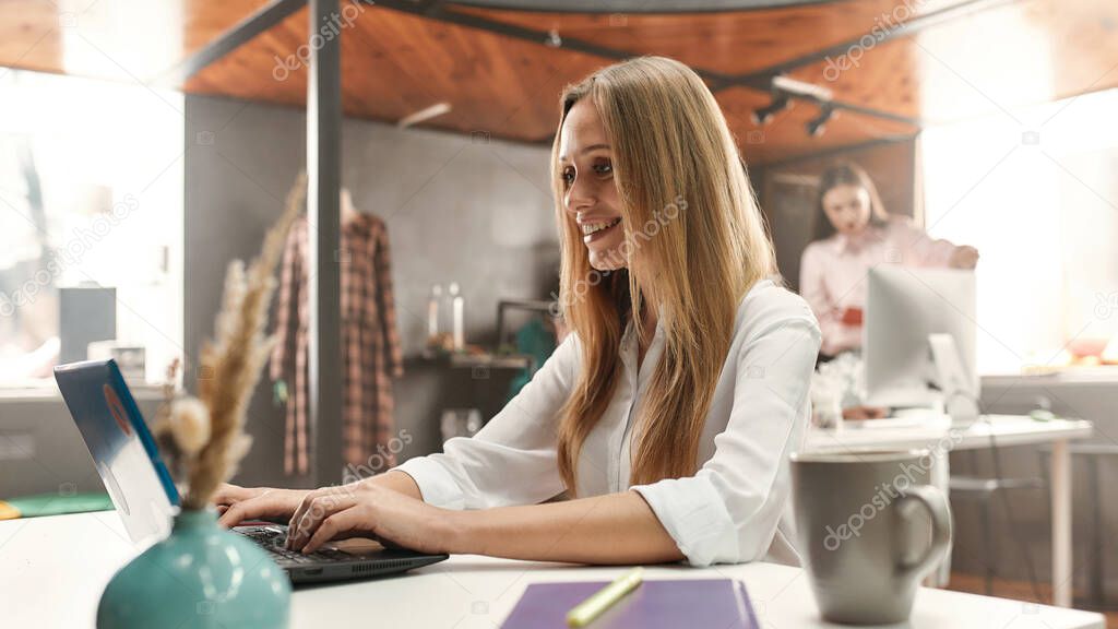 A pretty middle age woman with blond hair working with her laptop smiling while sitting at a table with her colleagues behind