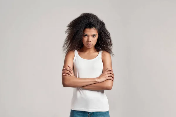 Ritratto di giovane donna afroamericana con i capelli ricci che indossa camicia bianca guardando focalizzata sulla macchina fotografica mentre in piedi con le braccia incrociate isolato su sfondo grigio — Foto Stock