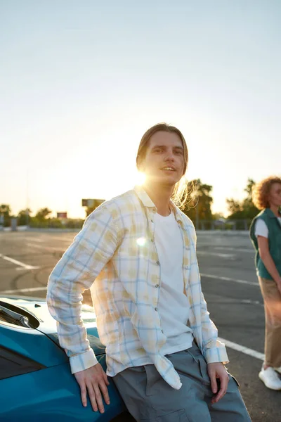 Een casual geklede witte langharige man kijkt in een camera leunend tegen een auto buiten op een parkeerplaats met zijn vrienden op een achtergrond — Stockfoto