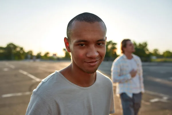 Um close-up de um homem de pele escura em uma camisa branca sorrindo de pé fora em um local de estacionamento com seu amigo em um fundo — Fotografia de Stock