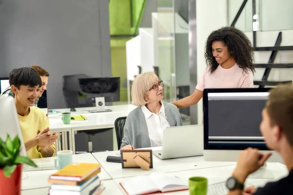 Idős nő, vezető gyakornok nézi a női kollégáját, miközben befejezi az első munkáját, barátságos munkások tapsolnak, éljenzik az új alkalmazottakat az irodában. — Stock Fotó