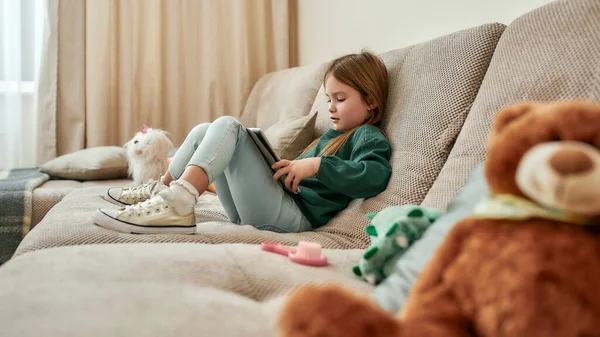 Una niña sentada en un sofá con sus zapatos viendo dibujos animados en una tableta —  Fotos de Stock