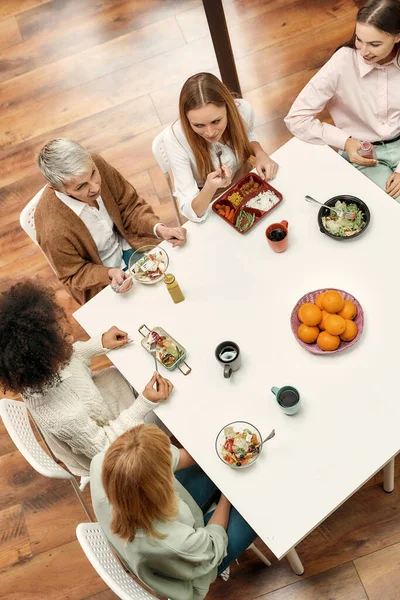 Eine Gruppe von fünf kreativen Frauen unterschiedlichen Alters sitzt gut gekleidet an einem Tisch und isst gesundes Essen und spricht miteinander — Stockfoto