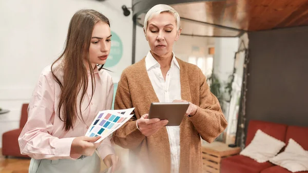 Um branco maduro bem vestido ladyboss segurando um tablet dando instruções para um jovem branco moda vestido senhora — Fotografia de Stock