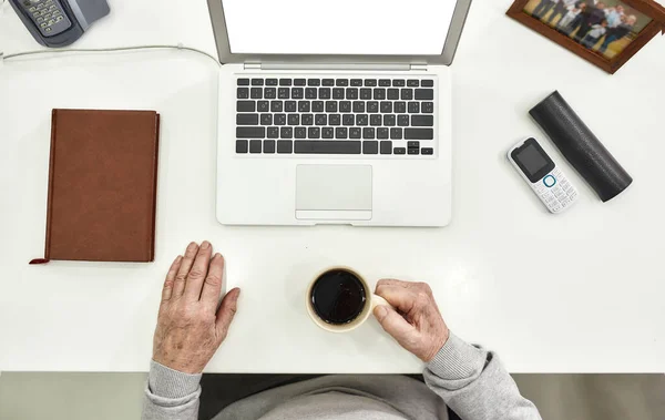 Top view of aged male employee senior intern holding a cup of coffee while sitting at desk in modern office, Notebook, picture frame, cell phone, glasses and laptop on the table — Stock Photo, Image