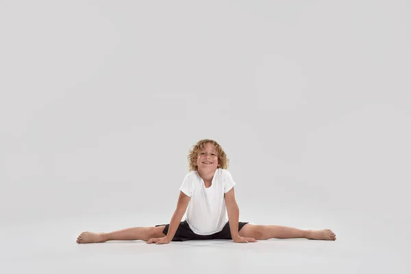 Full length shot of little playful boy child smiling at camera, doing splits isolated over grey background — Stock Photo, Image