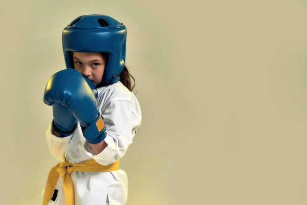 Portrait of little karate girl in white kimono wearing gloves and protective helmet, looking focused at camera, ready to punch, doing martial arts isolated over yellow background — Stock Photo, Image