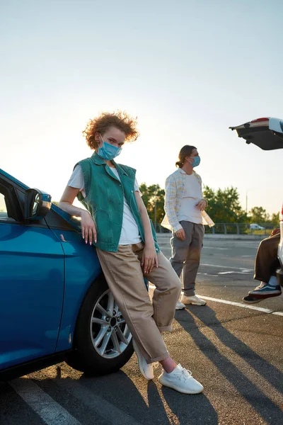 Een goed geklede roodharige jonge vrouw leunend tegen een auto met een masker in een camera die buiten op een parkeerplaats staat met haar vrienden op een achtergrond — Stockfoto