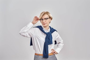 Şık orta yaşlı beyaz kadın portresi iş elbisesi ve gözlük takmış poz verirken kameraya bakıyor, gri arka planda tek başına duruyor.