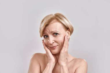 Hassas, duygusal, orta yaşlı bir kadının portresi kameraya bakıyor, yanaklarına dokunuyor, gri arka planda izole bir şekilde poz veriyor. Güzellik, cilt bakımı