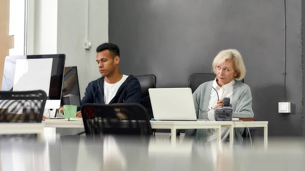 Aged woman, senior intern looking uncertain while using laptop, sitting at desk, working in modern office with other young employee — Stock Photo, Image