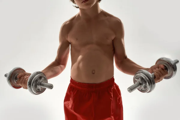 Cropped shot of little sportive boy child with muscular body lifting weights while standing isolated over white background — Stock Photo, Image