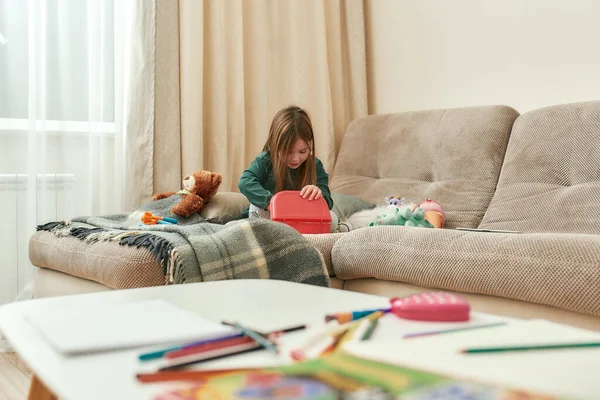 Una linda niña pequeña abriendo una pequeña caja de juguetes roja con juguetes sentados en un sofá en una habitación luminosa — Foto de Stock