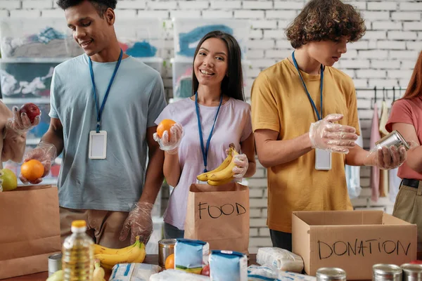 Joven voluntaria en guantes sonriendo a la cámara mientras empaca la donación de alimentos en bolsas de papel y caja, equipo pequeño que trabaja en la fundación benéfica — Foto de Stock