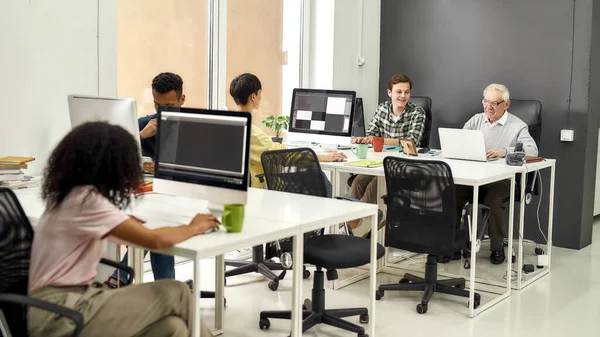 Полный снимок пожилого человека, старшего стажера, печатающего, используя ноутбук, сидя за столом, работая вместе с другими молодыми сотрудниками в современном офисе — стоковое фото