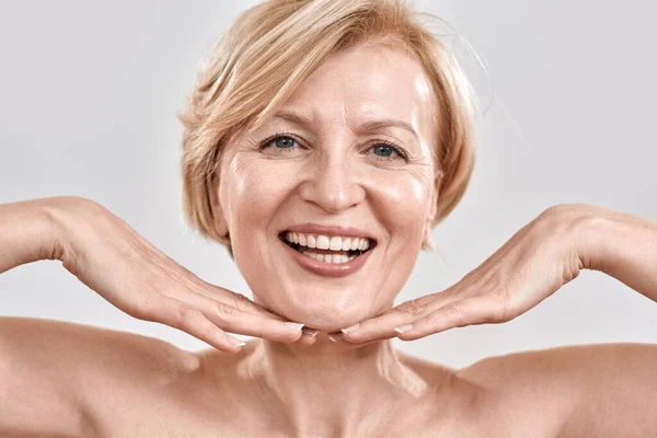 Close up retrato de bela mulher de meia-idade satisfeito com o tratamento da pele. Ela está segurando as mãos perto de seu rosto, sorrindo para a câmera isolada contra fundo cinza. Conceito de beleza — Fotografia de Stock