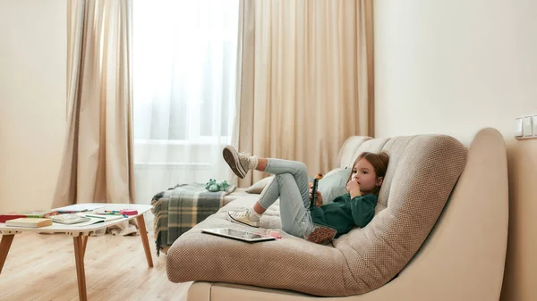 Uma menina bonita sentada sozinha perna a perna em um sofá assistindo TV em uma grande sala brilhante — Fotografia de Stock