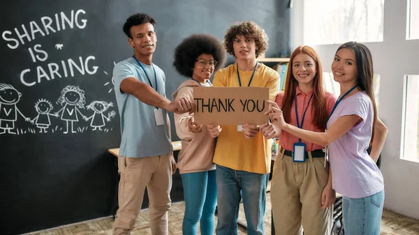 Group of diverse young volunteers smiling at camera, holding card with Thank you lettering while standing in charitable organization office