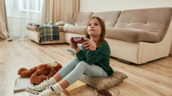 Una pequeña chica linda mira atentamente a una pantalla de un televisor jugando videojuego sosteniendo un mando mientras está sentada en el suelo de una habitación espaciosa —  Fotos de Stock