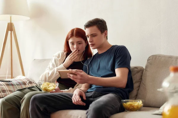 Un par de adolescentes sentados en el sofá en casa, mirando emocionalmente mientras miran algo por teléfono, usando el mismo par de auriculares —  Fotos de Stock