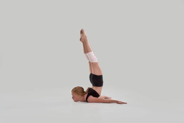 Full length shot of cute little redhead girl, professional gymnast raised her legs up, showing flexibility isolated over grey background — Stock Photo, Image