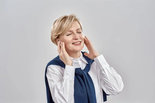 Retrato de una elegante mujer caucásica de mediana edad vestida con atuendo de negocios, ajustando sus auriculares, sonriendo con los ojos cerrados mientras posa aislada sobre un fondo gris —  Fotos de Stock