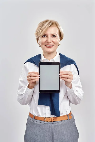 Ritratto di elegante donna caucasica di mezza età che indossa abiti da lavoro sorridente alla macchina fotografica, tenendo tablet pc con schermo vuoto, applicazione pubblicitaria mentre in piedi isolato su sfondo grigio — Foto Stock