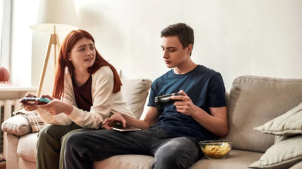 Adolescente mirando irritado a su novio mientras se distraía con su teléfono móvil. Pareja joven sosteniendo la consola de juegos, jugando videojuegos juntos en casa —  Fotos de Stock