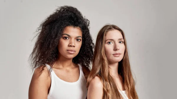 Ritratto ravvicinato di due giovani donne diverse che indossano camicie bianche guardando fiduciosi davanti alla macchina fotografica mentre posano insieme isolati su sfondo grigio — Foto Stock