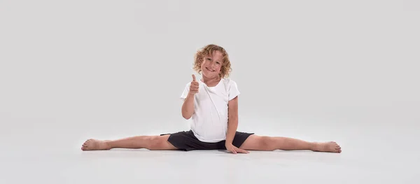 Full length shot of little playful boy child smiling at camera, showing thumbs up while doing splits isolated over grey background — Stock Photo, Image