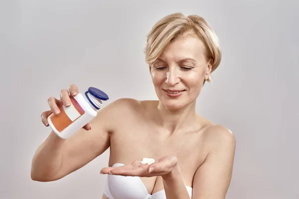 Portrait of middle aged caucasian woman pouring the pills out of the bottle while standing isolated over grey background. Healthcare concept — Stock Photo, Image