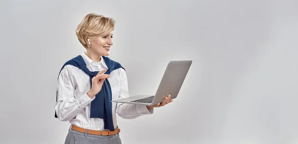 İş kıyafetleri içinde dizüstü bilgisayar giyen zarif orta yaşlı beyaz kadın portresi, çevrimiçi video görüşmesi yaparken, gri arka planda tek başına dikiliyordu. — Stok fotoğraf