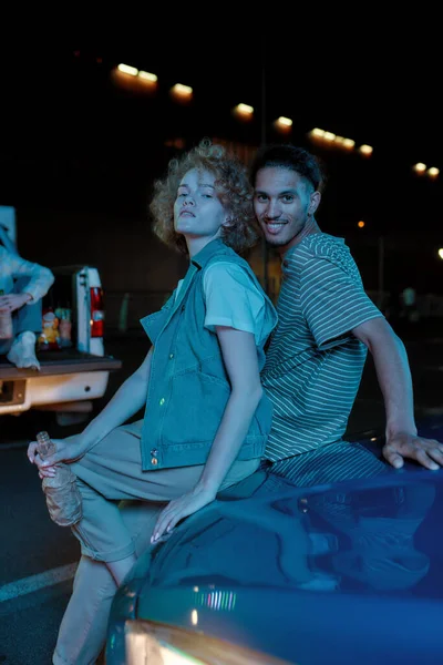 Hezký mladý bílý pár různých národností sedí na kufru auta na parkovišti a dívá se do kamery pózující a pijící pivo — Stock fotografie