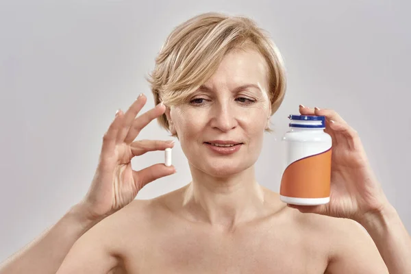 Potret wanita Kaukasia paruh baya yang memegang botol dengan pil dan pil di tangannya, melihatnya sambil berdiri terisolasi di atas latar belakang abu-abu. Konsep layanan kesehatan. — Stok Foto