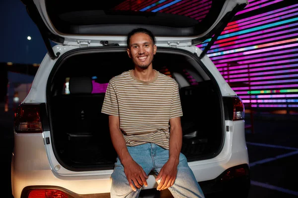 A well-dressed dark-skinned young latina man with a piercing smiling looking into a camera sitting in an opened car trunk outside on a parking site with a led screen behind