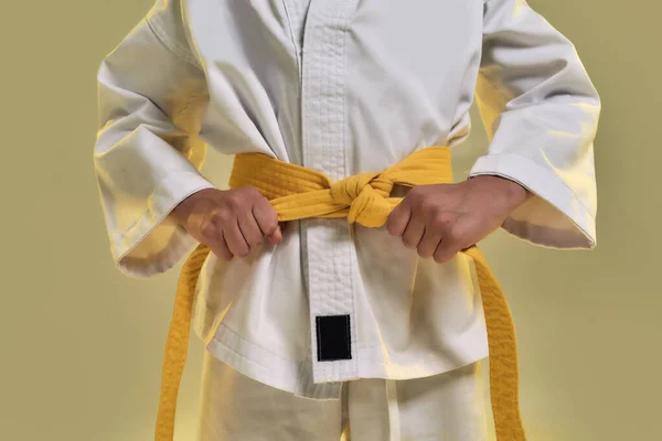 Beyaz kimonolu küçük karateci kızın egzersiz yapmaya hazırlanırken sarı kuşak ayarladığı, dövüş sanatları yaptığı, sarı arka planda tek başına durduğu sahneler. — Stok fotoğraf