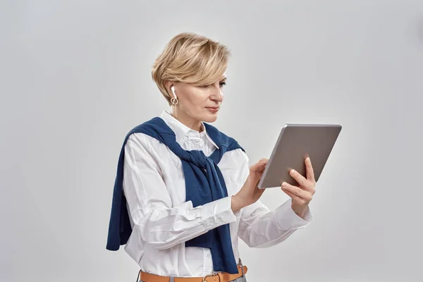 사업 복을 입고 이어폰을 들고 회색 배경에 고립 된 채 태블릿 PC 를 사용하는 중년의 우아 한 여성의 모습 — 스톡 사진