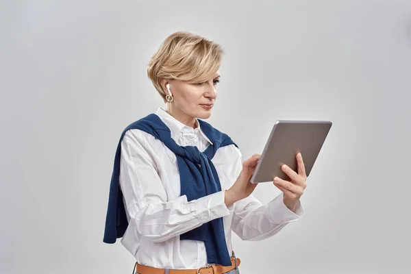 Ritratto di elegante donna caucasica di mezza età che indossa abiti da lavoro e auricolari in possesso, utilizzando tablet pc mentre in piedi isolato su sfondo grigio — Foto Stock