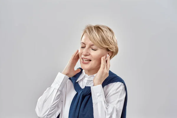 Retrato de mulher branca de meia-idade elegante vestindo trajes de negócios, ajustando seus fones de ouvido, sorrindo com os olhos fechados enquanto posando isolado sobre fundo cinza — Fotografia de Stock