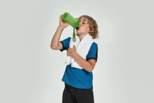Little sportive boy child in sportswear drinking water from the bottle while standing with towel around his neck isolated over white background — Stock Photo, Image
